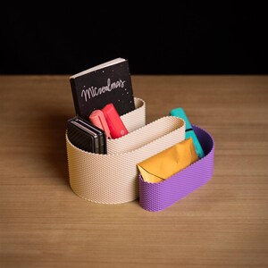 May include: A beige and purple desk organiser with a textured, grid-like surface. The organiser holds a black notebook with the text Microalmas, a black rectangular case, two pink pens, a turquoise marker, and a small yellow envelope.