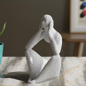 May include: A white abstract sculpture of a seated figure with a textured, granular surface. The figure is depicted in a contemplative pose with both hands raised to the sides of the head. The sculpture has a smooth, curved silhouette.