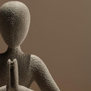 Może przedstawiać: Teksturowana, szara rzeźba przedstawiająca osobę w pozycji medytacyjnej. Postać ma gładką, owalną głowę i ramiona złożone do modlitwy. Rzeźba jest ustawiona na ciepłym, neutralnym tle.