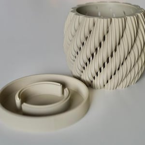 May include: A cream-colored, decorative planter with a unique, textured design. The planter features a spiral pattern with openings, and sits next to a matching, circular base. The design is modern and would complement any home decor.