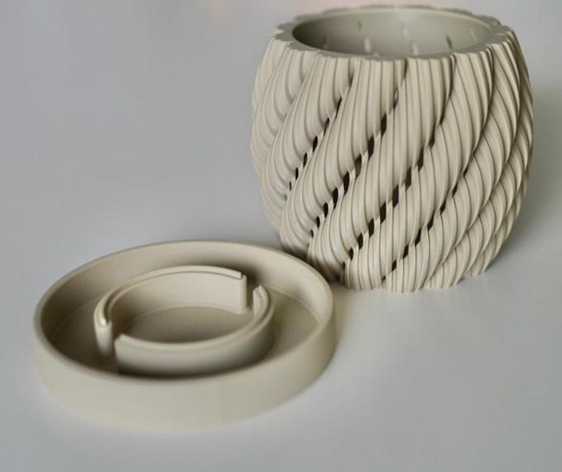 May include: A cream-colored, decorative planter with a unique, textured design. The planter features a spiral pattern with openings, and sits next to a matching, circular base. The design is modern and would complement any home decor.