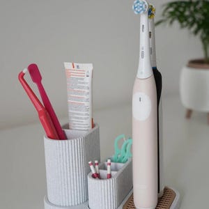 May include: A light gray toothbrush organizer with multiple compartments. It holds two pink toothbrushes, a tube of toothpaste, dental floss, and two electric toothbrushes. The organizer has a textured, ribbed design.