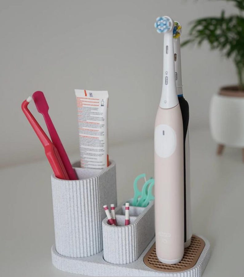 May include: A light gray toothbrush organizer with multiple compartments. It holds two pink toothbrushes, a tube of toothpaste, dental floss, and two electric toothbrushes. The organizer has a textured, ribbed design.