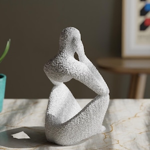 May include: A white sculpture of a seated figure with a textured, granular surface. The figure is depicted in a contemplative pose with one arm resting on a knee and the hand touching the face, creating a hollow space within the abstract form.