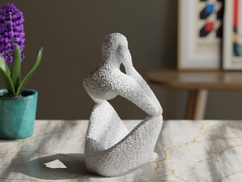 May include: A white sculpture of a seated figure with a textured, granular surface. The figure is depicted in a contemplative pose with one arm resting on a knee and the hand touching the face, creating a hollow space within the abstract form.