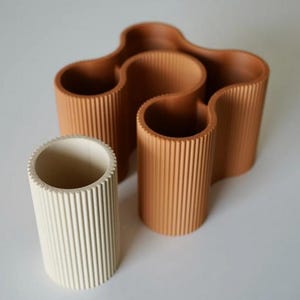 May include: A collection of three decorative vases. One is off-white, cylindrical, and the other two are a warm brown color. The brown vases have a wavy design and a ribbed texture. The vases are made of a solid material and are suitable for home decor.