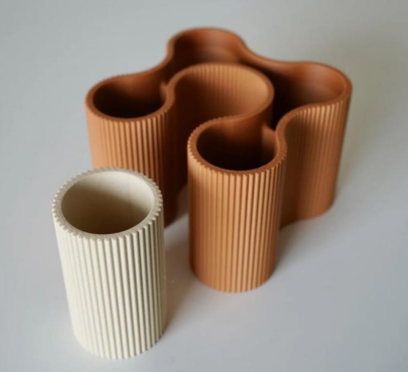May include: A collection of three decorative vases. One is off-white, cylindrical, and the other two are a warm brown color. The brown vases have a wavy design and a ribbed texture. The vases are made of a solid material and are suitable for home decor.