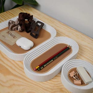 May include: A set of three white and speckled oval trays with a textured brown interior. The trays contain items such as pencils, hair accessories, a white earbud case, and binder clips. The word "SIMIU" is visible on a card.