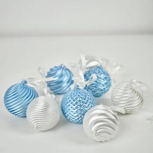 Pode incluir: Uma cole&ccedil;&atilde;o de ornamentos esf&eacute;ricos em tons de azul claro e prateado. Alguns ornamentos apresentam uma textura canelada em espiral, enquanto outros exibem um padr&atilde;o de grade em losango. Cada ornamento tem uma pequena fita branca para pendurar.