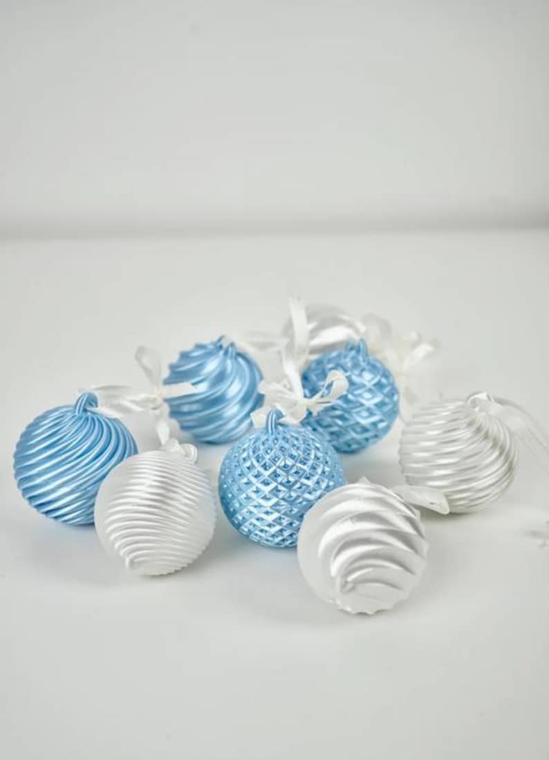 Pode incluir: Uma cole&ccedil;&atilde;o de ornamentos esf&eacute;ricos em tons de azul claro e prateado. Alguns ornamentos apresentam uma textura canelada em espiral, enquanto outros exibem um padr&atilde;o de grade em losango. Cada ornamento tem uma pequena fita branca para pendurar.