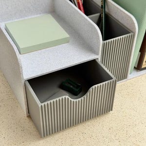 Op de afbeelding: Een lichtgrijze gespikkelde bureau-organizer met een uitschuifbare lade met een verticaal geribbelde voorkant, een open bovencompartiment met lichtgroene plakbriefjes en een achtergedeelte met verdelers voor pennen en potloden.