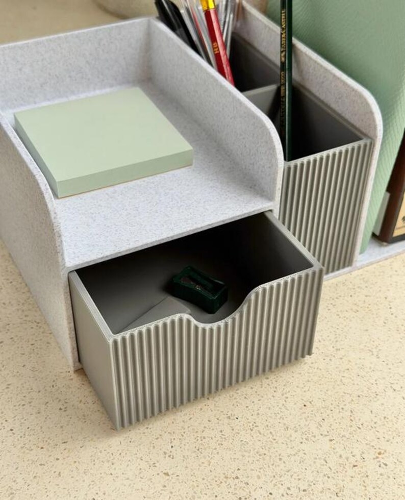 Op de afbeelding: Een lichtgrijze gespikkelde bureau-organizer met een uitschuifbare lade met een verticaal geribbelde voorkant, een open bovencompartiment met lichtgroene plakbriefjes en een achtergedeelte met verdelers voor pennen en potloden.
