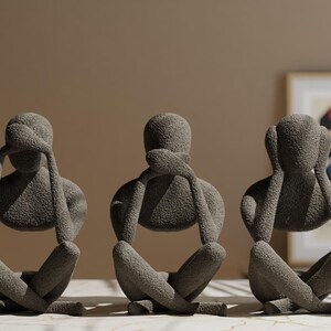 K&ouml;nnte beinhalten: Drei graue Skulpturen in sitzender Pose, jede mit H&auml;nden, die Augen, Ohren und Mund bedecken. Die Figuren haben eine strukturierte Oberfl&auml;che und sind auf einer gemusterten Oberfl&auml;che angeordnet. Der Hintergrund ist neutral gef&auml;rbt.