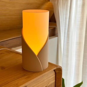 May include: A cylindrical table lamp with a matte beige exterior shell that curves upward to reveal a glowing, warm orange light source. The lamp features a smooth, petal-like overlapping design that encases the inner light, creating a soft illumination.