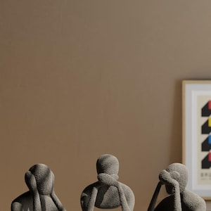 K&ouml;nnte beinhalten: Drei graue abstrakte Skulpturen von sitzenden Figuren, jede in einer anderen Pose, die ihre Augen, Ohren oder ihren Mund bedecken. Die Figuren stehen auf einer marmor&auml;hnlichen Oberfl&auml;che mit neutralem braunem Hintergrund.