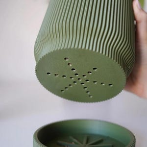 May include: A green, ribbed plant pot with a matching saucer. The pot has a unique, wavy design and drainage holes in the bottom. The saucer has a star-shaped design in the center. The pot and saucer are made of a matte material.