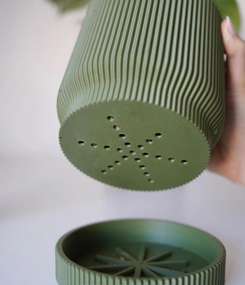 May include: A green, ribbed plant pot with a matching saucer. The pot has a unique, wavy design and drainage holes in the bottom. The saucer has a star-shaped design in the center. The pot and saucer are made of a matte material.