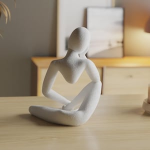 May include: A white abstract sculpture of a seated figure with a textured surface. The figure is depicted in a contemplative pose with one arm resting on a knee and the head tilted, showcasing smooth, flowing lines and a solid, matte white finish.