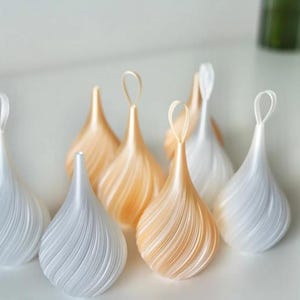 May include: A collection of teardrop-shaped ornaments featuring a spiraling ribbed texture. The ornaments are finished in alternating shades of pearlescent white and soft champagne gold, each topped with a matching fabric loop for hanging.