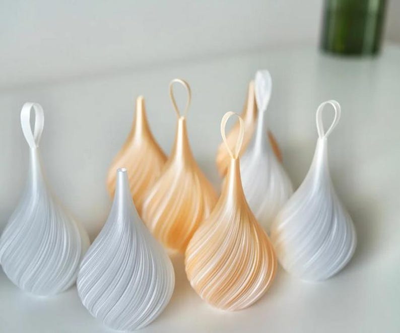 May include: A collection of teardrop-shaped ornaments featuring a spiraling ribbed texture. The ornaments are finished in alternating shades of pearlescent white and soft champagne gold, each topped with a matching fabric loop for hanging.