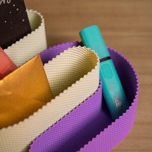 May include: A desk organiser featuring flexible, wavy-edged walls in purple and cream colours. The organiser holds a turquoise highlighter, a gold envelope, and a black card with white text. The walls have a fine, grid-like textured surface.