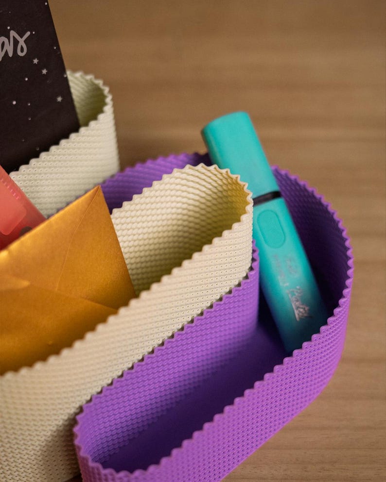 May include: A desk organiser featuring flexible, wavy-edged walls in purple and cream colours. The organiser holds a turquoise highlighter, a gold envelope, and a black card with white text. The walls have a fine, grid-like textured surface.