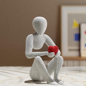 Puede incluir: Una figura gris texturizada de una persona sentada sosteniendo un coraz&oacute;n rojo. La escultura tiene un dise&ntilde;o minimalista, con una superficie rugosa y moteada. El coraz&oacute;n es de un rojo vibrante, contrastando con la figura gris. La figura es un objeto decorativo.