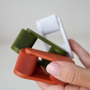 May include: Three colorful, handheld lint rollers in shades of orange, green, and white. The rollers have a ribbed texture and are held in a person's hand. The background is a soft, neutral color.
