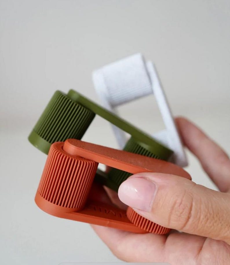 May include: Three colorful, handheld lint rollers in shades of orange, green, and white. The rollers have a ribbed texture and are held in a person's hand. The background is a soft, neutral color.