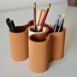 May include: A terracotta-colored desk organizer with a wavy design. The organizer has multiple cylindrical compartments holding pencils, pens, and markers. The pencils have pink erasers and yellow accents. The pens and markers are black and cream-colored.