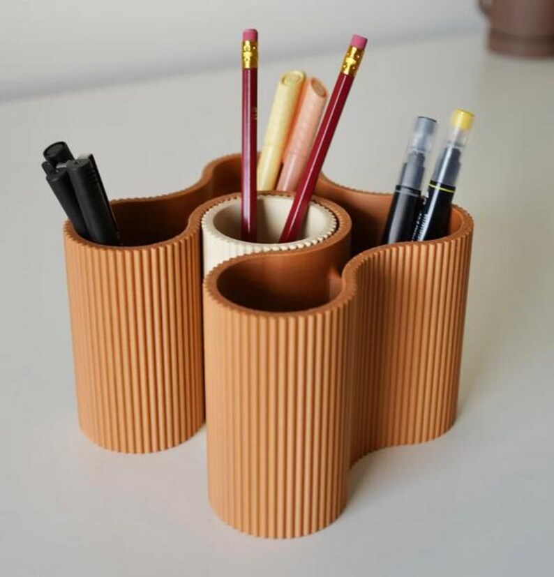 May include: A terracotta-colored desk organizer with a wavy design. The organizer has multiple cylindrical compartments holding pencils, pens, and markers. The pencils have pink erasers and yellow accents. The pens and markers are black and cream-colored.