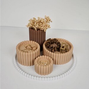 May include: A collection of three beige fluted cylindrical storage containers, a dark brown fluted vase filled with dried cream-colored flowers, a brown scrunchie, a brown hair clip, and gold-toned jewelry, all arranged on a white beaded tray.