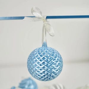 Pode incluir: Um ornamento esf&eacute;rico azul-claro com uma superf&iacute;cie texturizada de padr&atilde;o de diamante. O ornamento pende de uma fita de cetim branca fina atada num la&ccedil;o no topo. A superf&iacute;cie apresenta um design de grelha geom&eacute;trica repetitivo.