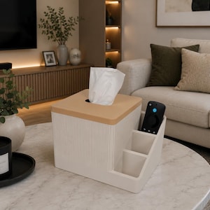 Puede incluir: Un soporte rectangular para cajas de pañuelos de color blanco roto con textura acanalada y tapa de color tostado. La unidad cuenta con un compartimento de almacenamiento integrado con dos ranuras verticales, una con un mando negro y un pañuelo blanco.
