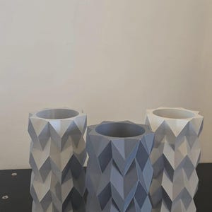 Puede incluir: Tres jarrones geom&eacute;tricos en tonos grises y blancos. Cada jarr&oacute;n tiene forma cil&iacute;ndrica con un patr&oacute;n en zigzag. Los jarrones est&aacute;n colocados sobre una superficie negra contra un fondo neutro.