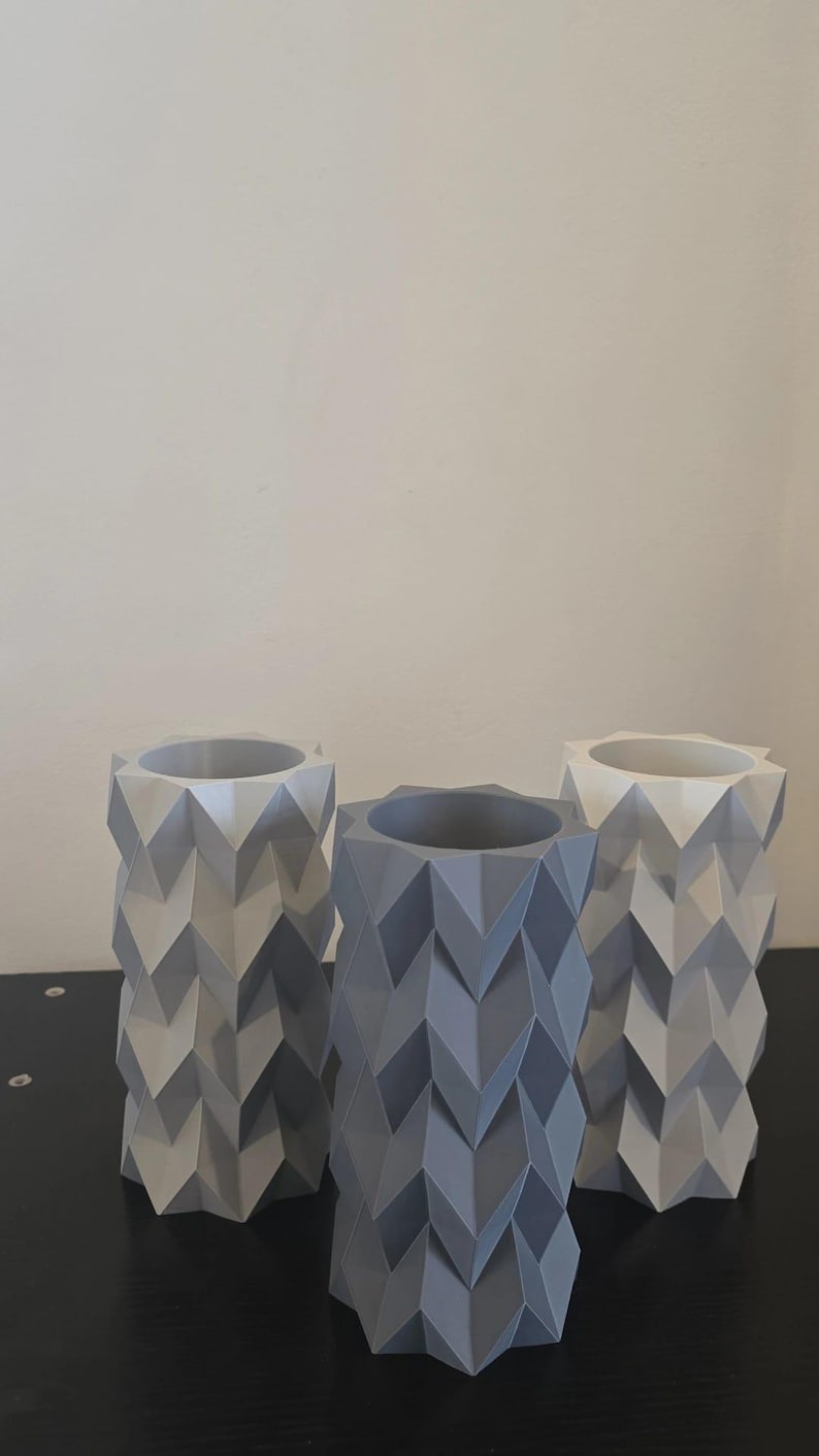 Puede incluir: Tres jarrones geom&eacute;tricos en tonos grises y blancos. Cada jarr&oacute;n tiene forma cil&iacute;ndrica con un patr&oacute;n en zigzag. Los jarrones est&aacute;n colocados sobre una superficie negra contra un fondo neutro.