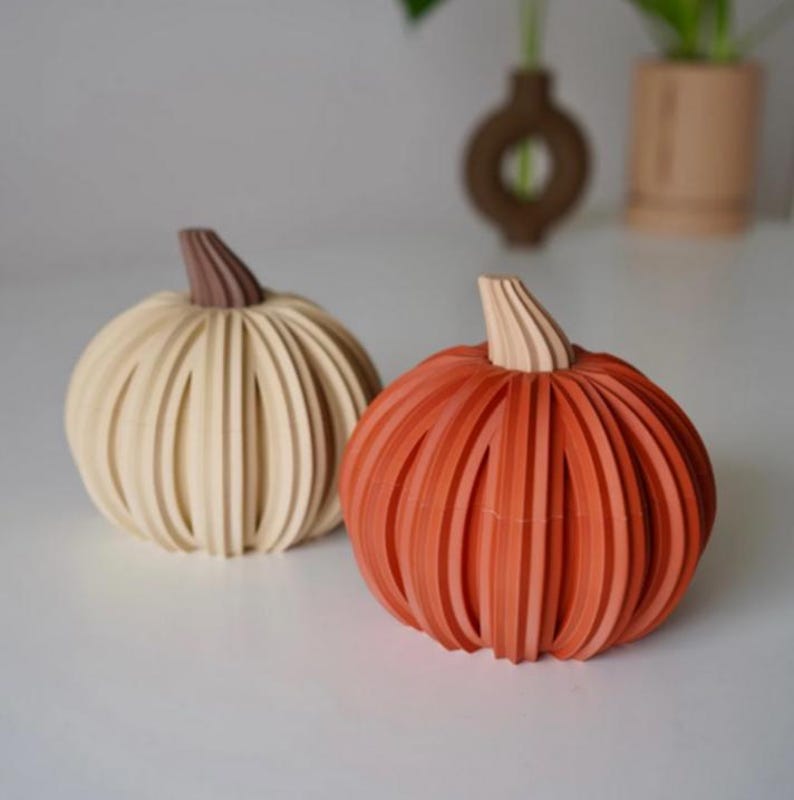 May include: Two decorative pumpkins with a ribbed, layered design. One pumpkin is cream-colored with a brown stem, and the other is orange with a light beige stem. Both objects feature a rounded shape with vertical ridges throughout the exterior.