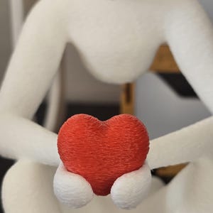 Puede incluir: Una figura blanca de una persona sentada con las rodillas dobladas, sosteniendo un coraz&oacute;n rojo texturizado. El coraz&oacute;n es el punto focal, con la forma de la figura borrosa en el fondo. La figura tiene una apariencia suave y texturizada.
