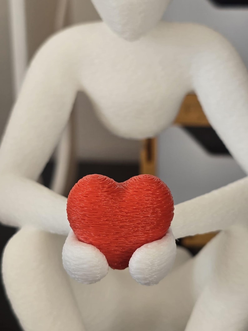 Puede incluir: Una figura blanca de una persona sentada con las rodillas dobladas, sosteniendo un coraz&oacute;n rojo texturizado. El coraz&oacute;n es el punto focal, con la forma de la figura borrosa en el fondo. La figura tiene una apariencia suave y texturizada.