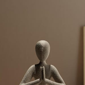 Może przedstawiać: Szara, teksturowana rzeźba przedstawiająca osobę w pozycji siedzącej jogi ze złączonymi dłońmi w modlitwie. Postać ma gładką, zaokrągloną głowę i siedzi ze skrzyżowanymi nogami. Rzeźba ma neutralny kolor i matowe wykończenie.