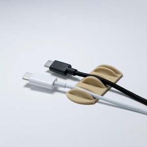 May include: A beige cable organiser with three curved slots holding two USB-C cables. One cable is white with a smooth finish, and the other is black with a braided texture. The organiser has a low-profile, rectangular base with rounded edges.