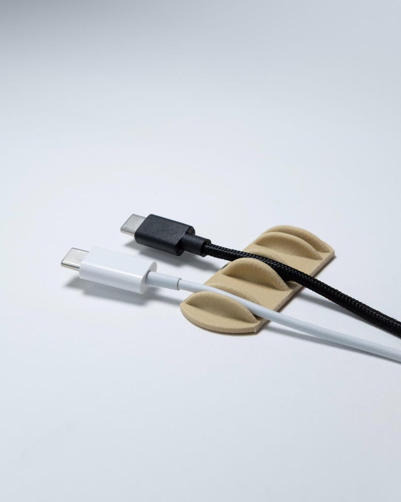 May include: A beige cable organiser with three curved slots holding two USB-C cables. One cable is white with a smooth finish, and the other is black with a braided texture. The organiser has a low-profile, rectangular base with rounded edges.