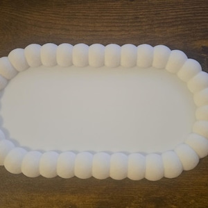 May include: A white, oval-shaped tray with a beaded edge. The tray's interior is smooth and flat, while the border features a series of rounded, textured beads. The tray is designed for serving or display.