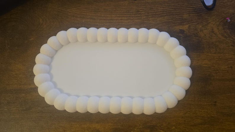 May include: A white, oval-shaped tray with a beaded edge. The tray's interior is smooth and flat, while the border features a series of rounded, textured beads. The tray is designed for serving or display.