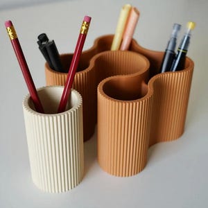 May include: A cream-colored pencil holder and a wavy, tan-colored desk organizer with vertical ribbed detailing. The pencil holder contains two red pencils. The desk organizer holds pens and markers, providing a stylish storage solution for office supplies.