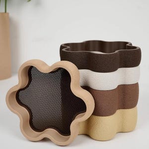 Puede incluir: Una pila de cuatro recipientes con forma de flor en tonos marr&oacute;n, beige y blanco. Un recipiente est&aacute; separado, revelando una malla. Los recipientes tienen una superficie texturizada.