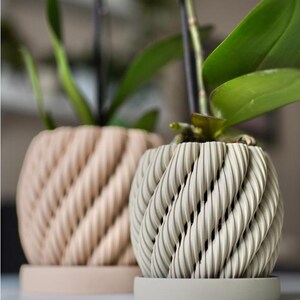 May include: Two decorative planters with a textured, woven design. One planter is a light beige color, and the other is a light gray color. Both planters have a matching round base. Green plant leaves are visible in the planters.