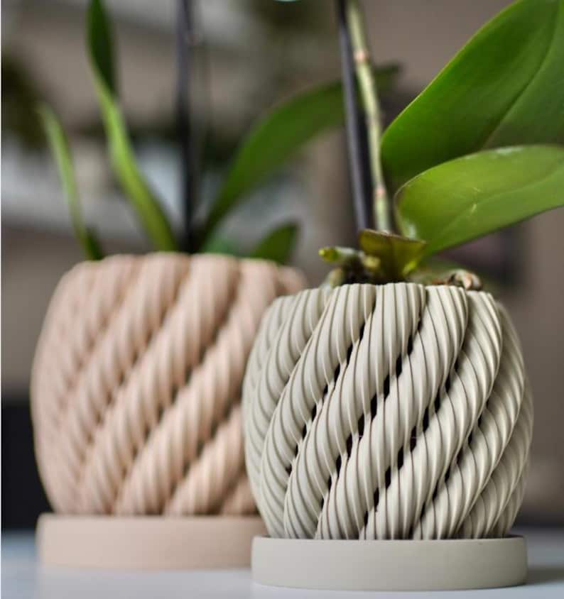 May include: Two decorative planters with a textured, woven design. One planter is a light beige color, and the other is a light gray color. Both planters have a matching round base. Green plant leaves are visible in the planters.