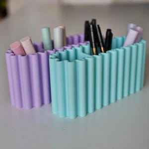 K&ouml;nnte beinhalten: Zwei pastellfarbene Beh&auml;lter mit gewelltem Rand. Einer ist lavendelfarben, der andere hellt&uuml;rkis. Die Beh&auml;lter enthalten verschiedene Make-up-Artikel und Stifte. Die Beh&auml;lter bestehen aus einem festen Material und stehen auf einer hellgrauen Oberfl&auml;che.
