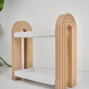 May include: A two-tiered decorative shelf in a neutral color palette. The shelf features a light beige frame with a rounded arch design and two white speckled shelves. The design is modern and minimalist, suitable for displaying small items.